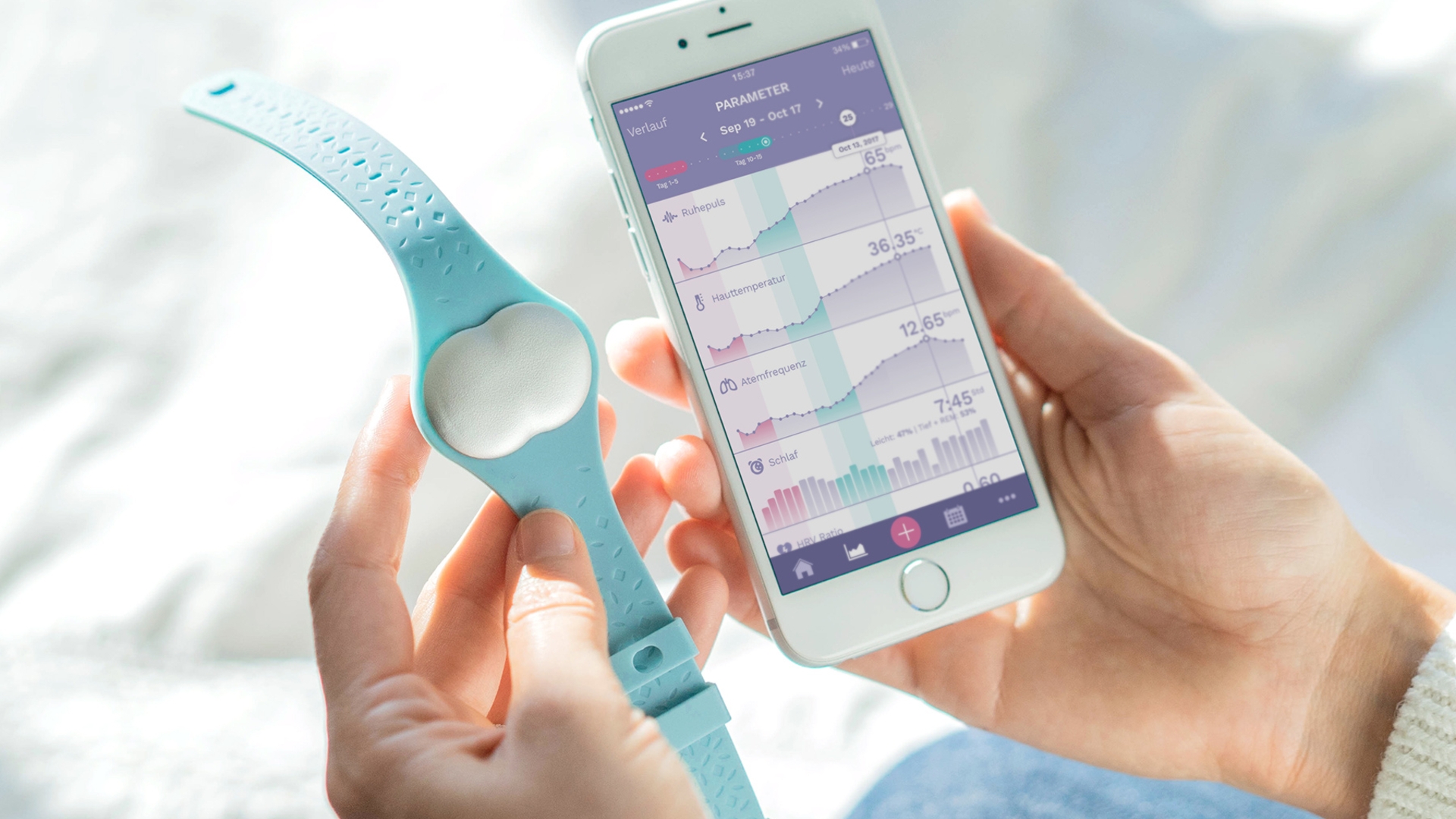 Ava Wearable Eine Frauenhand hält den babyblauen Fruchtbarkeitstracker Ava und die andere hält das Smartphone mit der Ava-App