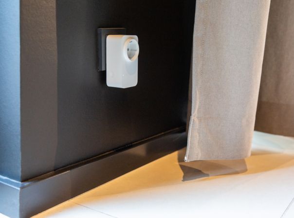 Amazon Smart Plug Eine Steckdose mit einem Steckdosenadapater in einer schwarzen Wand, daneben ein weißer Vorhang.