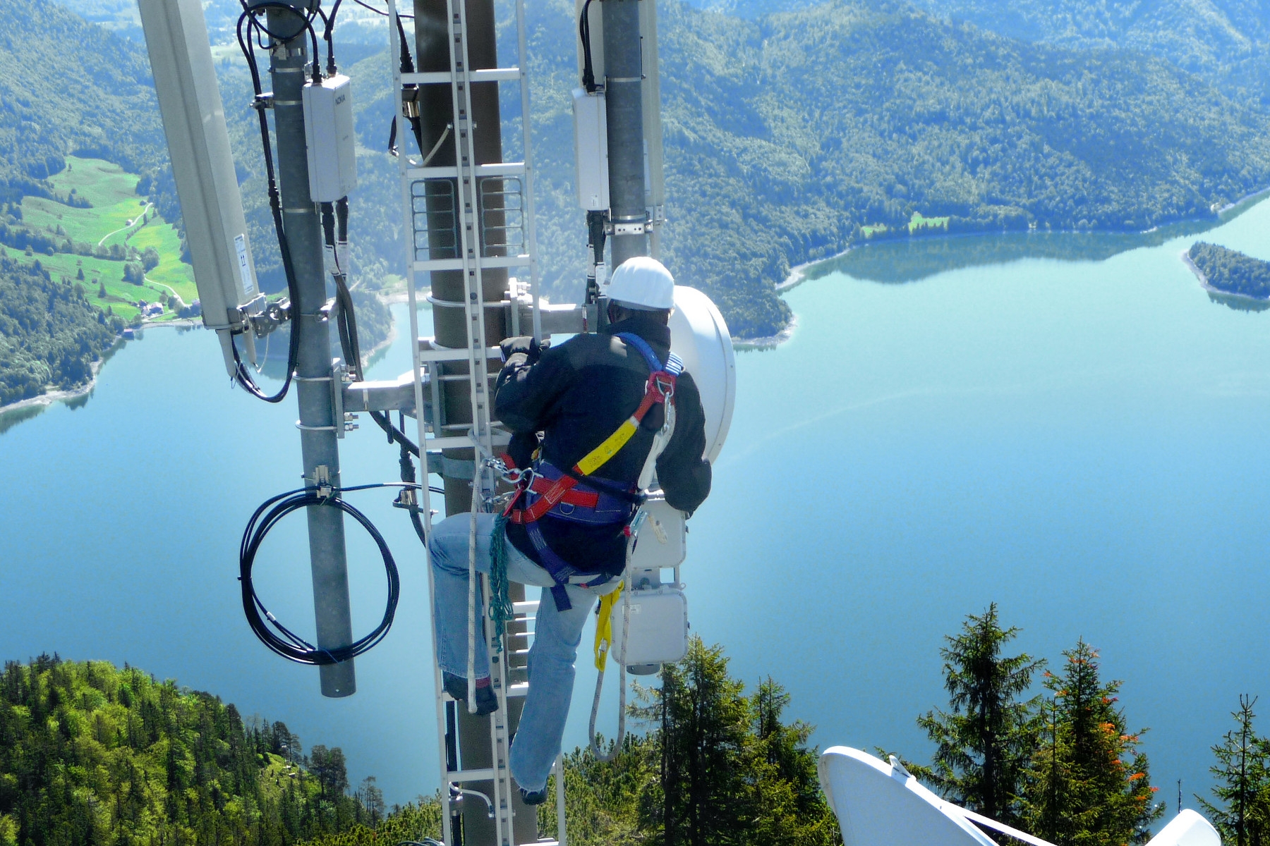 Techniker arbeitet in großer Höhe an Funkmasten vor Bergsee-Panorama